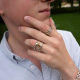 A person wearing glasses and a light blue shirt touches their chin, showcasing yellow gold annulets alongside the SPINELLI KILCOLLIN Phantom SK Ring and a chain bracelet. Green grass and trees are blurred in the background.