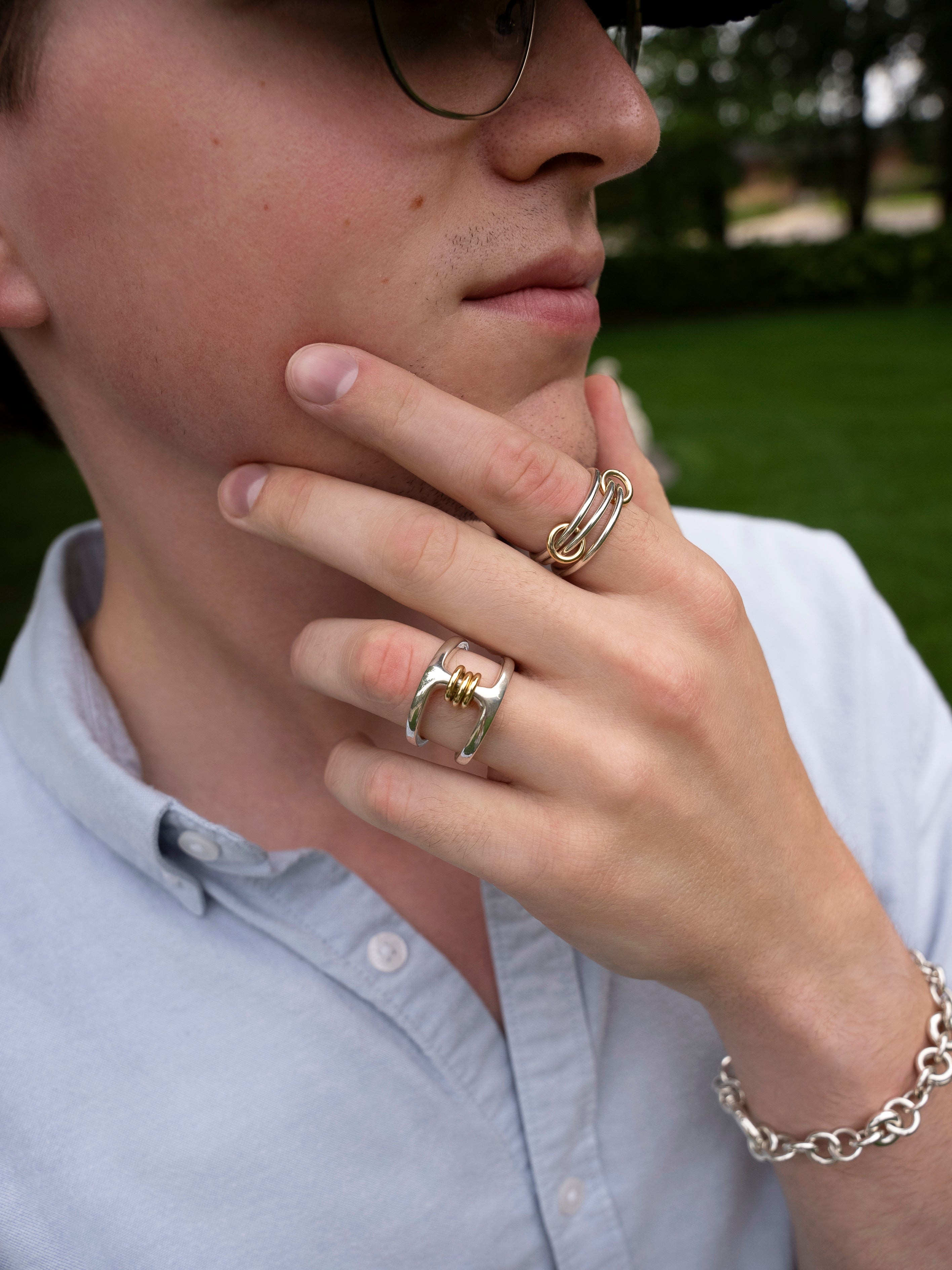 A person wearing glasses and a light blue shirt touches their chin, showcasing yellow gold annulets alongside the SPINELLI KILCOLLIN Phantom SK Ring and a chain bracelet. Green grass and trees are blurred in the background.