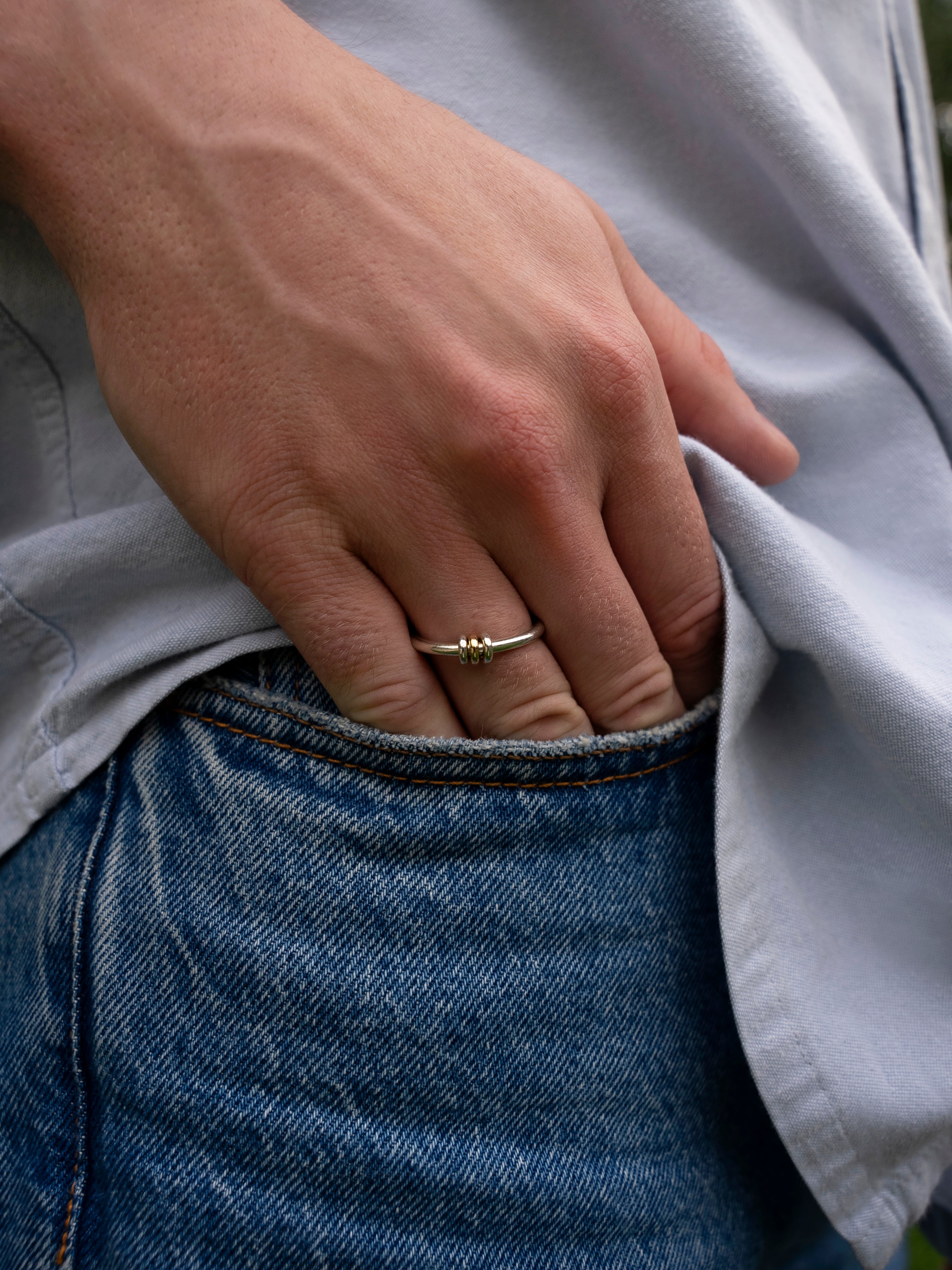 A close-up of a hand wearing the SPINELLI KILCOLLIN Sirius 3 Annulets Ring—a unisex gold ring with three beads—resting on the edge of a blue jean pocket. The person is also wearing a light gray shirt.
