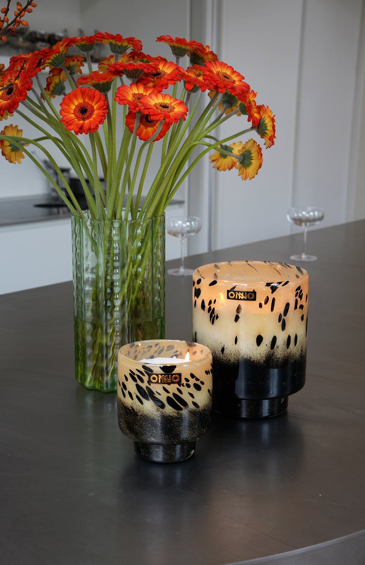 An ONNO Art Brigitte M Zanzibar Candle sits beside a green glass vase with orange and yellow flowers on a gray countertop, set against the backdrop of a modern, minimal kitchen.