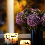 Two lit ONNO Art Brigitte S Zanzibar candles, featuring a chic black-and-white pattern, sit on a dark table. In the softly glowing light, a blurred vase of purple hydrangeas adds to the cozy, inviting atmosphere.