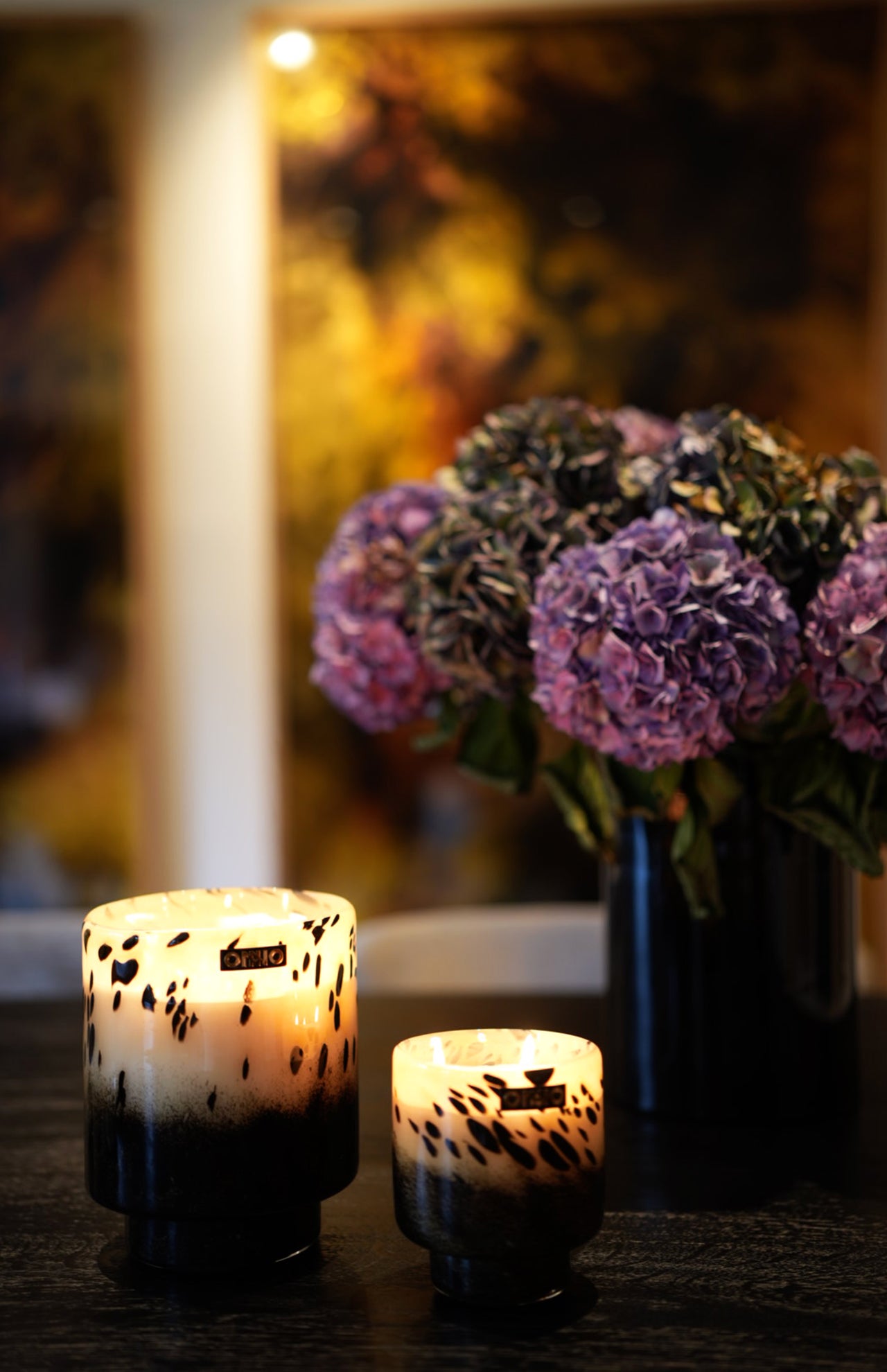 Two lit ONNO Art Brigitte S Zanzibar candles, featuring a chic black-and-white pattern, sit on a dark table. In the softly glowing light, a blurred vase of purple hydrangeas adds to the cozy, inviting atmosphere.