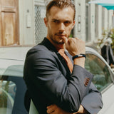 A man in a dark blazer leans against a light car outdoors, sunlight and buildings behind him. Thoughtfully crossing his arms, he wears the TATEOSSIAN Giza Double Click Pelle Bracelet, a navy braided leather accessory.