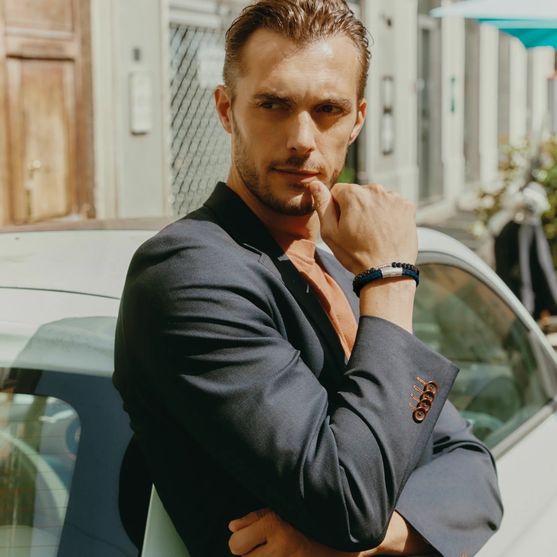 A man in a dark blazer leans against a light car outdoors, sunlight and buildings behind him. Thoughtfully crossing his arms, he wears the TATEOSSIAN Giza Double Click Pelle Bracelet, a navy braided leather accessory.