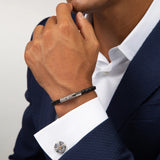 A man in a navy textured suit and white shirt showcases the TATEOSSIAN Vintage Herringbone Click Bracelet—a black braided leather piece with a stainless steel clasp—on his wrist, paired with a decorative cufflink for a refined look.