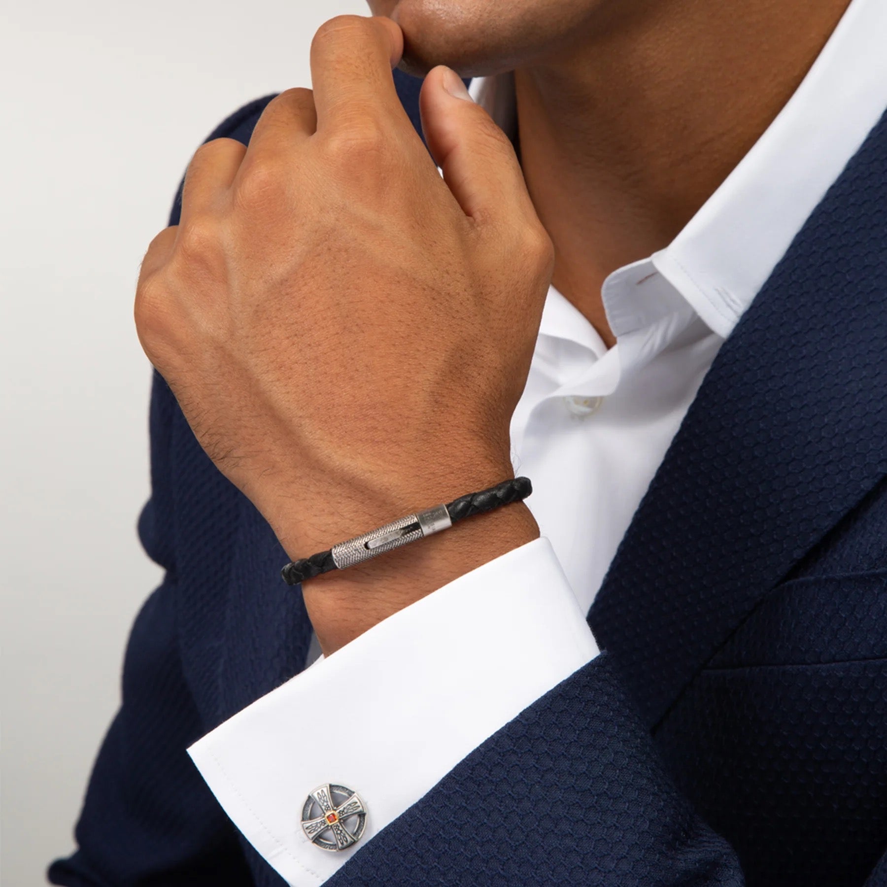 A man in a navy textured suit and white shirt showcases the TATEOSSIAN Vintage Herringbone Click Bracelet—a black braided leather piece with a stainless steel clasp—on his wrist, paired with a decorative cufflink for a refined look.