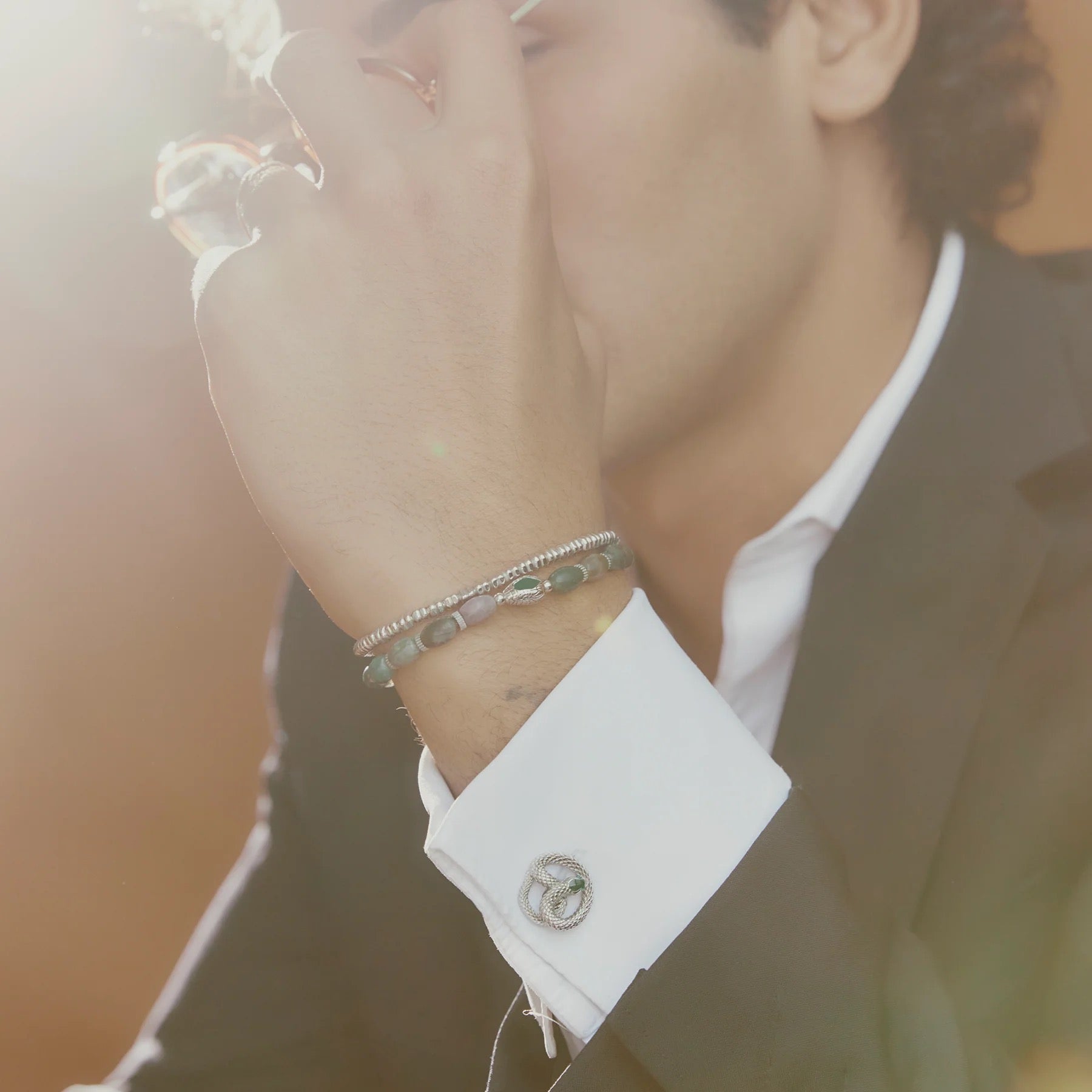 A suited individual poses with a TATEOSSIAN Indian Jasper Viper Bracelet on their wrist, paired with an elegant cufflink on their white shirt, as soft sunlight highlights the luxury accessories.
