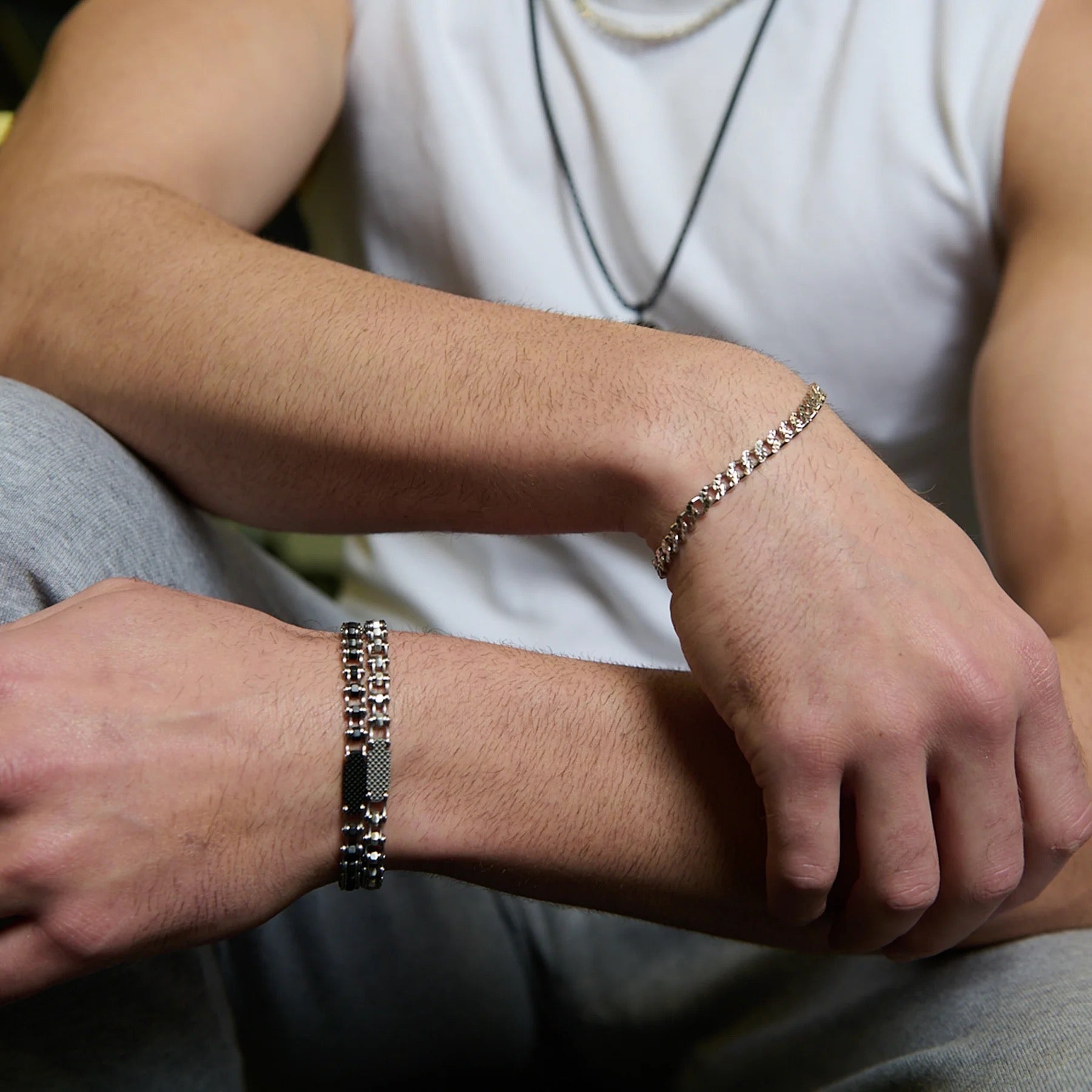 Wearing a sleeveless white shirt and gray pants, the person crosses their arms, showcasing two bracelets: TATEOSSIAN’s Bici Chain Bracelet on one wrist and a chunky stainless steel bracelet on the other.
