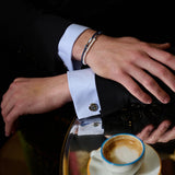 A person in a dark suit rests their hands on a reflective table, showing TATEOSSIAN cufflinks, a pearl bracelet, and the Feng Shui Fish Macrame Bracelet by TATEOSSIAN. An espresso cup with some coffee remains is on the table.