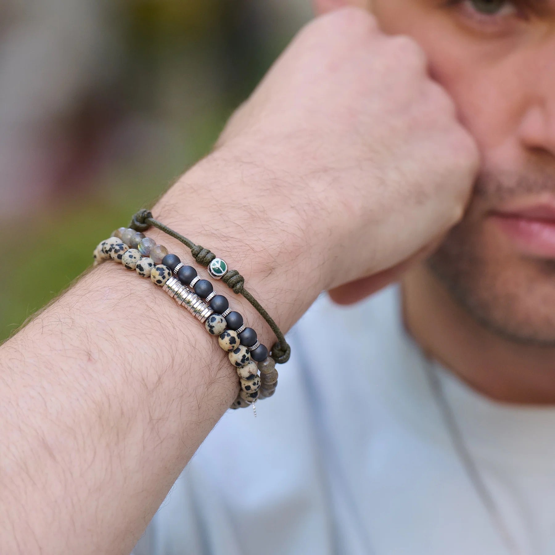 A close-up of a man's arm shows three bracelets: TATEOSSIAN's Classic Discs Dalmatian Jasper Bracelet, a sterling silver and black beaded bracelet, and a green string charm bracelet—stylishly made in London.