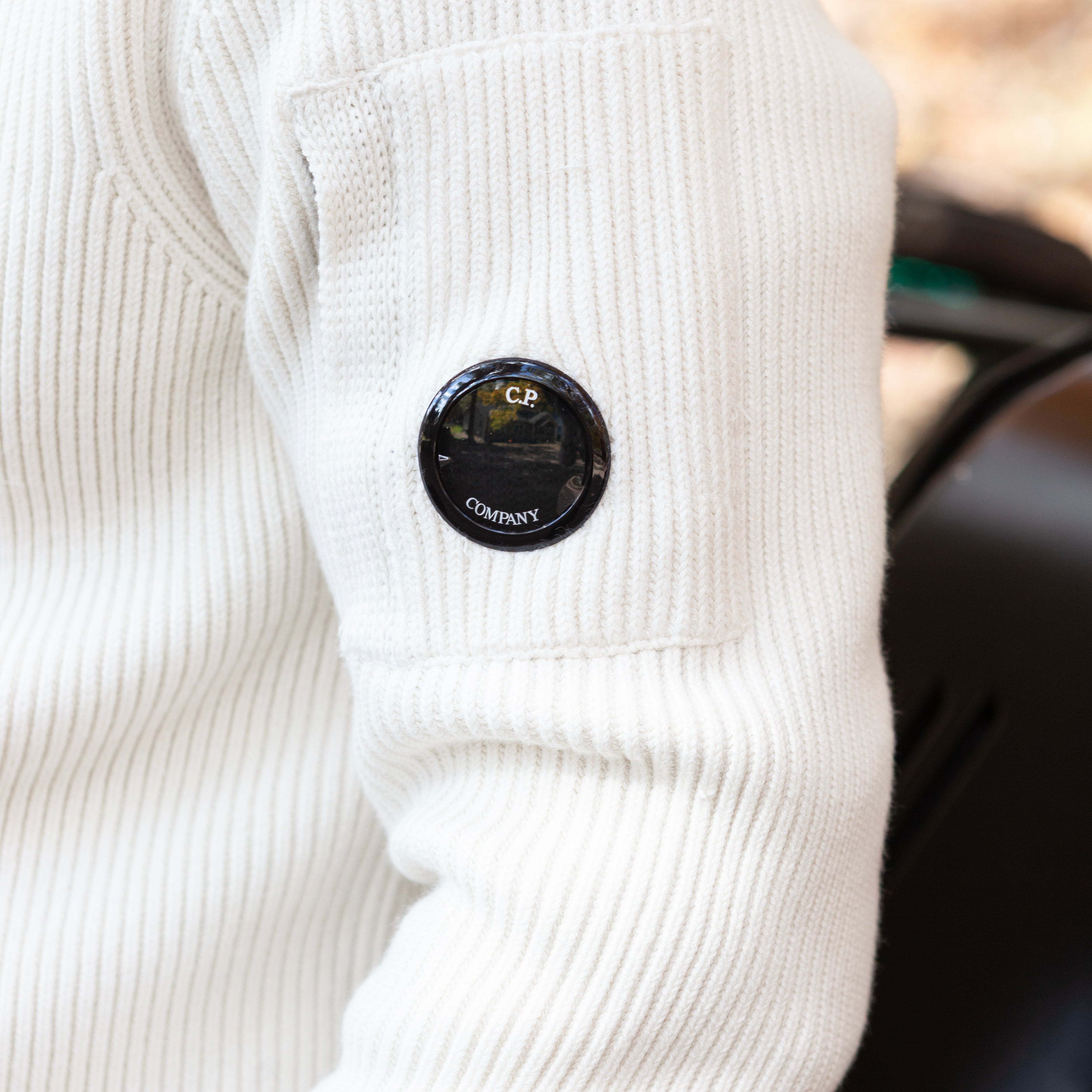 Close-up of a white ribbed sweater with a black circular logo on the chest.