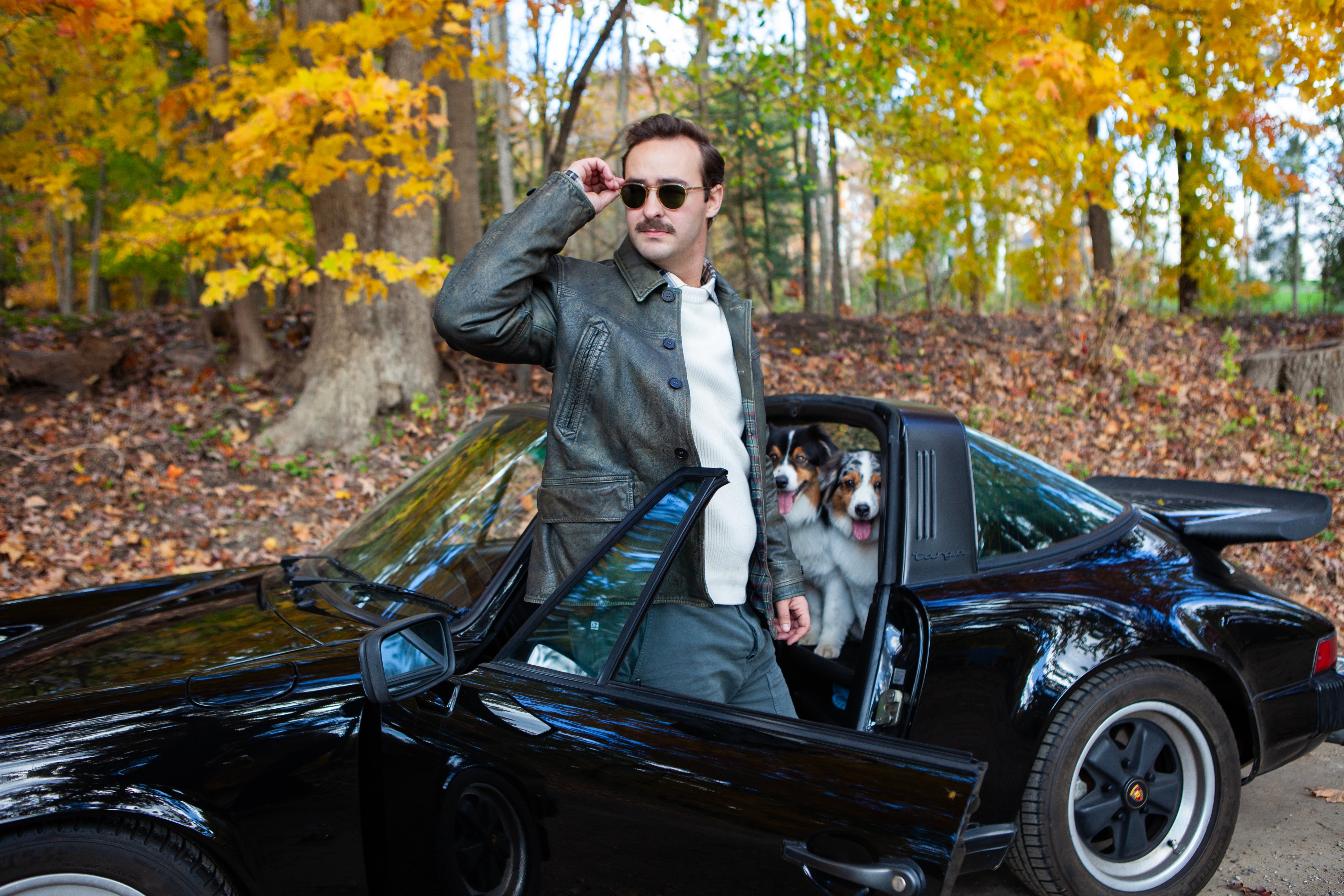 Man in a suit with sunglasses standing next to a black convertible car with a dog inside, autumn trees in the background.