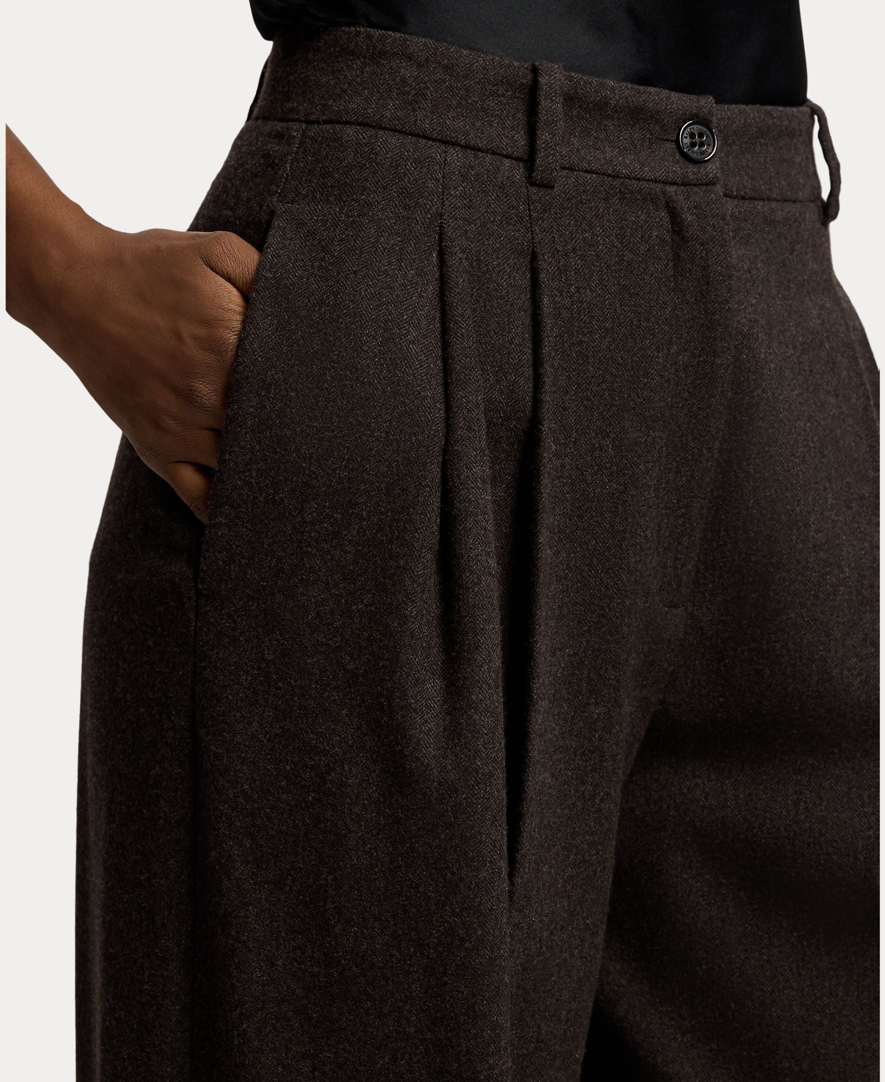 A person wearing the RALPH LAUREN COLLECTION Mini Herringbone Standard Pants with one hand in their pocket, showing the waistband, button, and part of a black shirt tucked in.