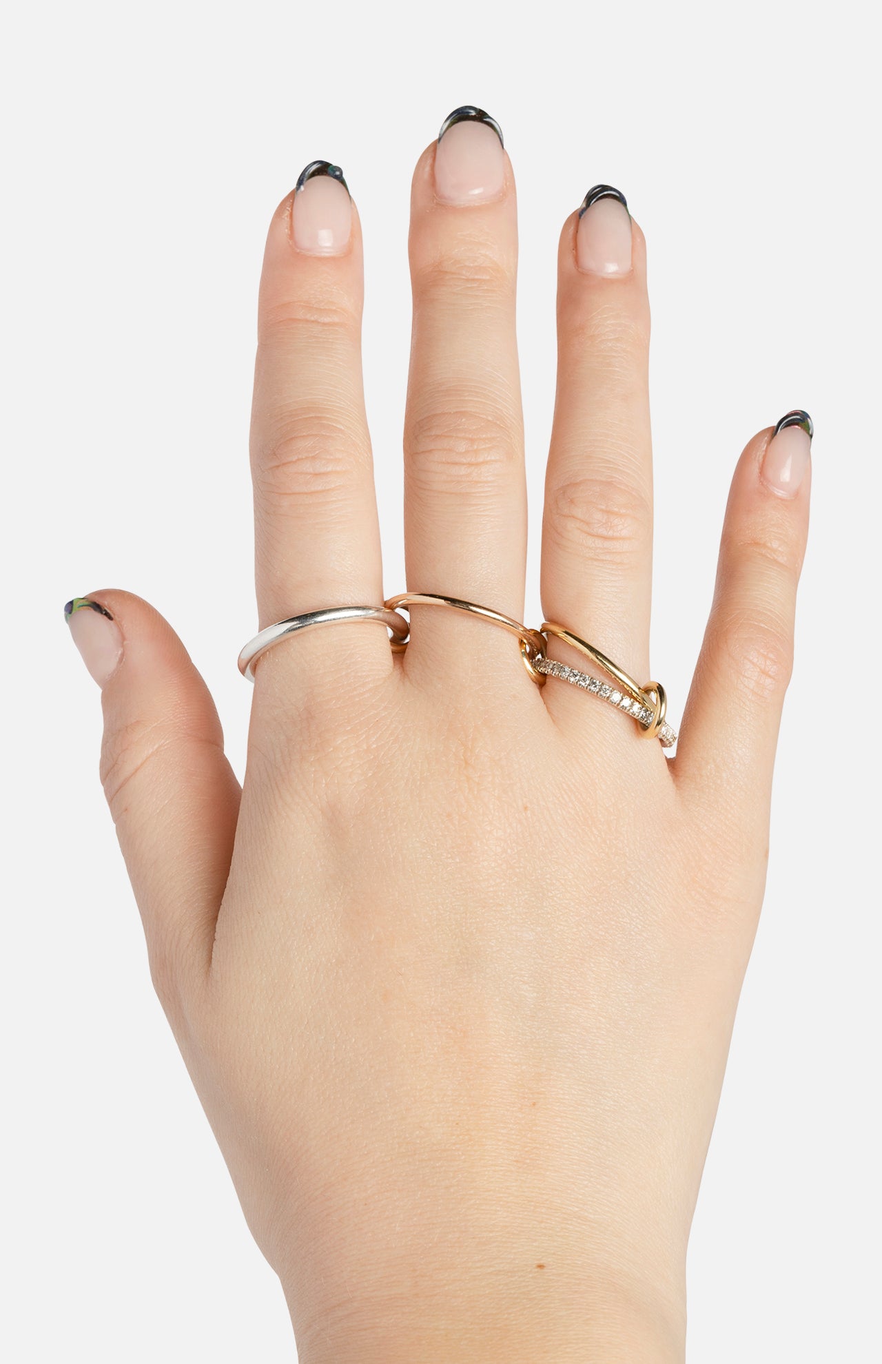 A hand with light skin and black-tipped French manicure nails wears the SPINELLI KILCOLLIN Nimbus 4 Band Diamond Ring along with two plain rings—one silver, one gold—against a white background.