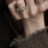 A close-up of a light-skinned person wearing the SPINELLI KILCOLLIN Raneth 3 Band Ring, hand resting on their chin. They have natural lips and wear a dark, textured jacket with frayed edges and visible stitching.