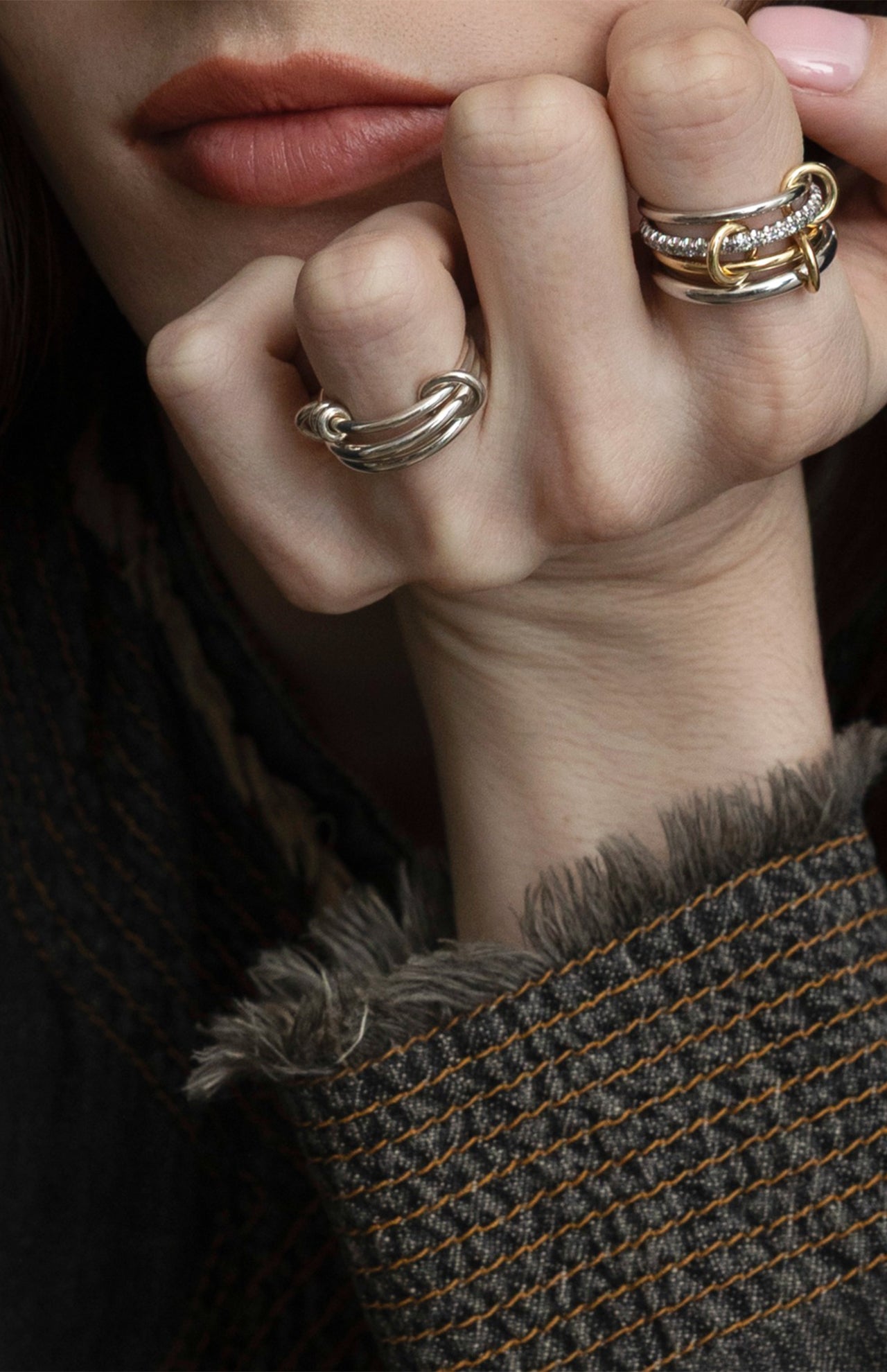 A close-up of a light-skinned person wearing the SPINELLI KILCOLLIN Raneth 3 Band Ring, hand resting on their chin. They have natural lips and wear a dark, textured jacket with frayed edges and visible stitching.
