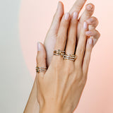 Two hands with manicured nails are elegantly posed, each adorned with stacked SPINELLI KILCOLLIN Raneth 3 Band Rings in fluid, modular gold designs. The soft background glows with warm pinkish light.