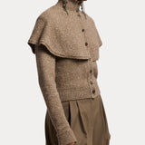 A person wears the RALPH LAUREN COLLECTION Cashmere Turtleneck Cardigan Sweater—a taupe, textured knit piece crafted in Italy—with its elegant capelet overlay, high ribbed turtleneck collar, long sleeves, and button-front design. The look is paired with loose brown trousers and finished with bold statement earrings.