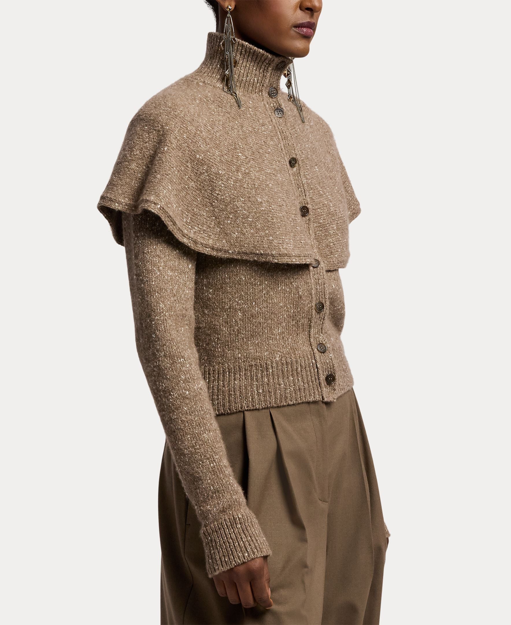 A person wears the RALPH LAUREN COLLECTION Cashmere Turtleneck Cardigan Sweater—a taupe, textured knit piece crafted in Italy—with its elegant capelet overlay, high ribbed turtleneck collar, long sleeves, and button-front design. The look is paired with loose brown trousers and finished with bold statement earrings.