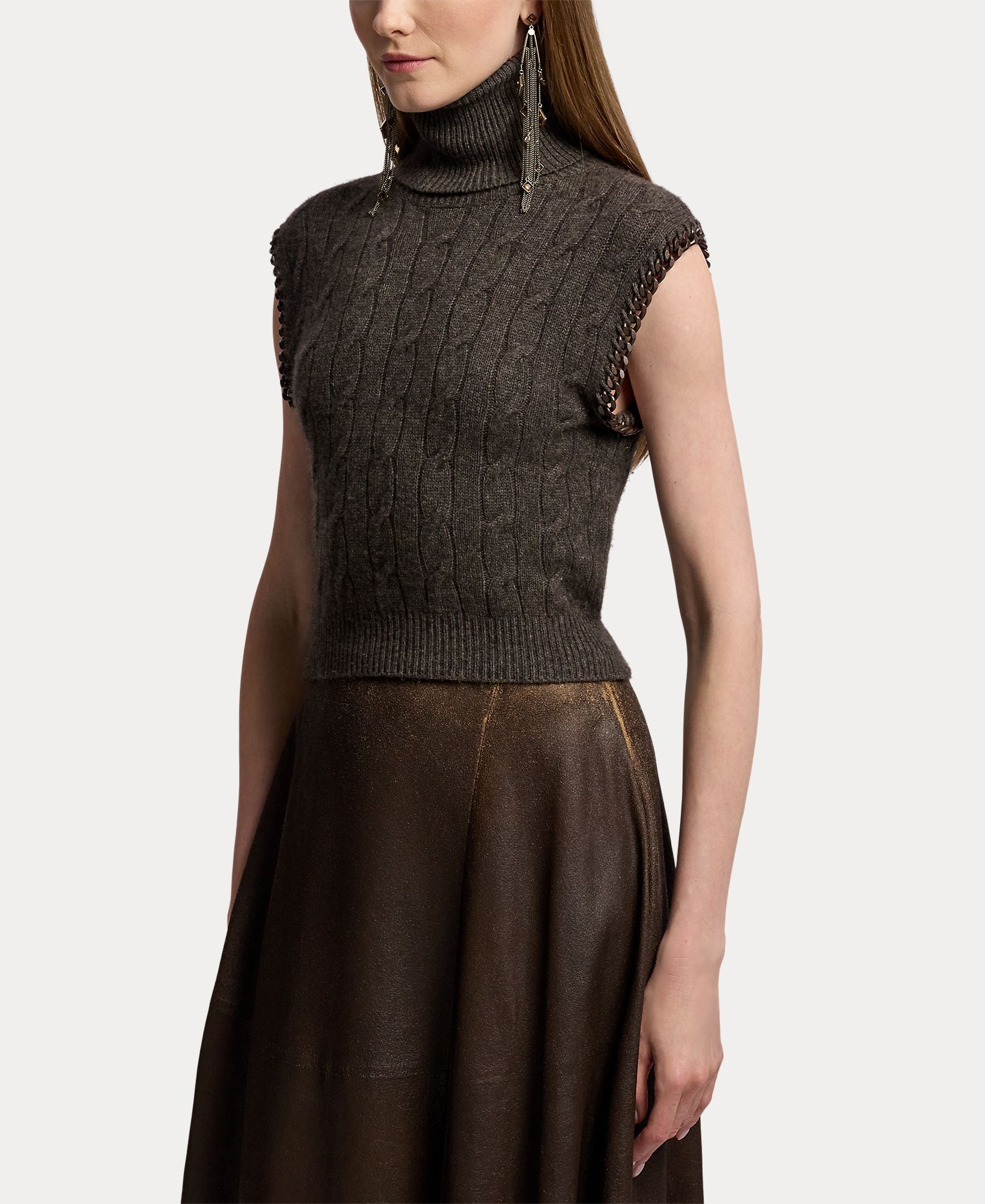A woman models the RALPH LAUREN COLLECTION Cashmere Turtleneck Sleeveless Sweater in dark gray, paired with a brown textured A-line skirt and long dangling earrings, against a plain light background.