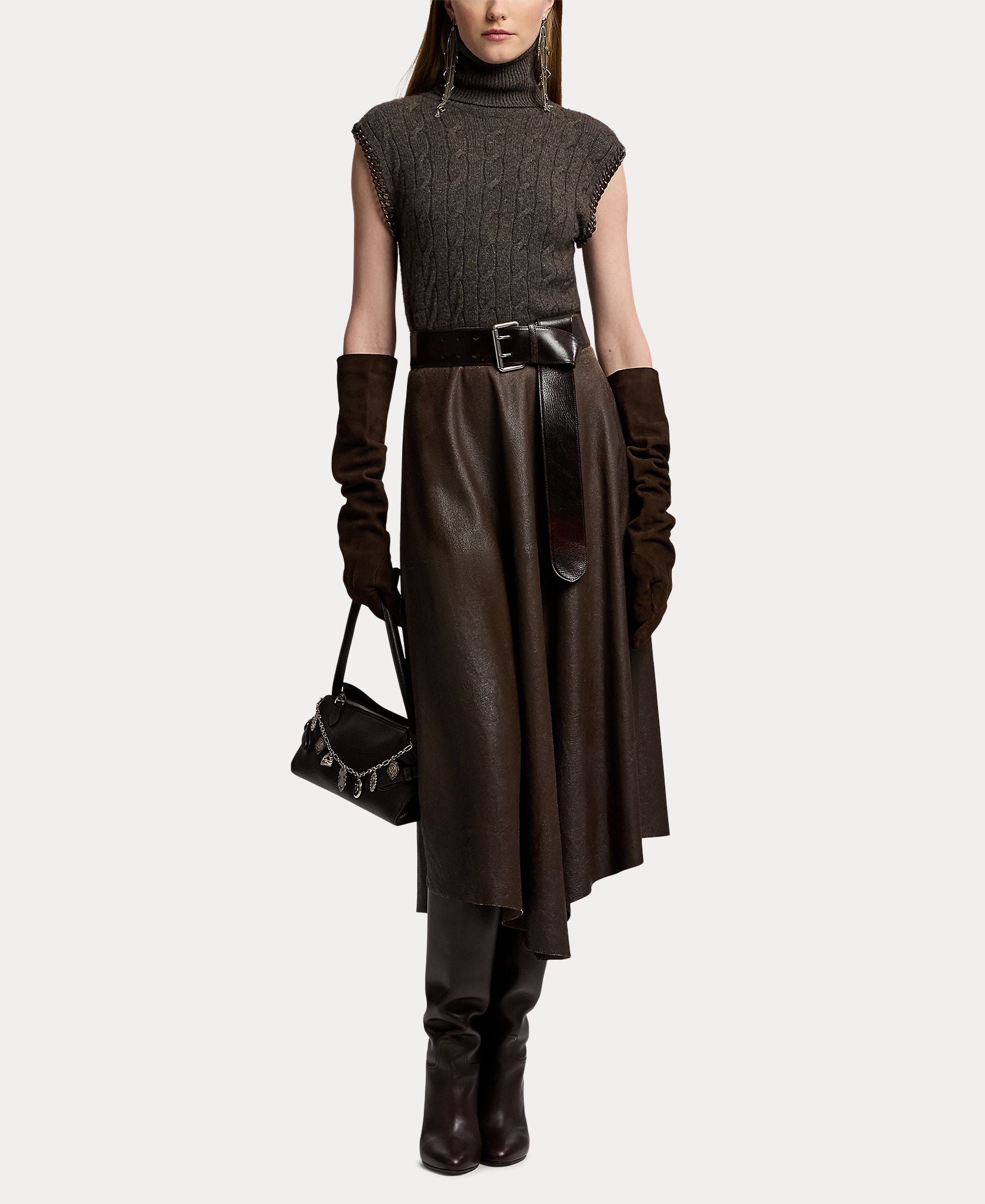 A woman wears the RALPH LAUREN COLLECTION Cashmere Turtleneck Sleeveless Sweater, paired with a belted midi skirt, long leather gloves, knee-high boots, and carries a small black handbag. The ensemble features dark tones for a sophisticated look against a white background.