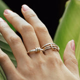 A hand with neatly manicured nails rests on a large green leaf, showcasing the SPINELLI KILCOLLIN Sirius 3 Annulets Ring—an elegant unisex sterling silver piece with intertwined minimalist designs, set against a softly blurred background.