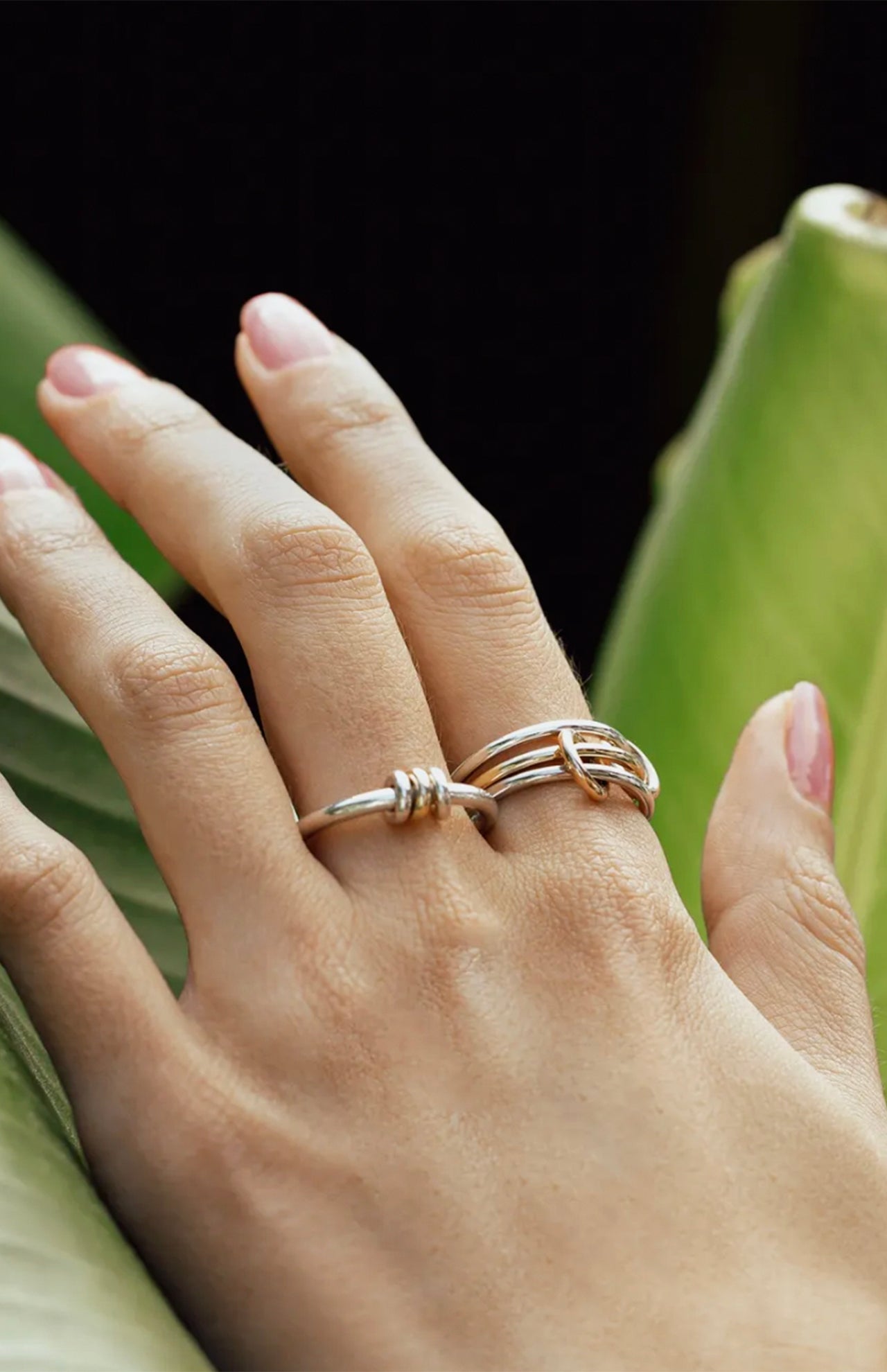 A hand with neatly manicured nails rests on a large green leaf, showcasing the SPINELLI KILCOLLIN Sirius 3 Annulets Ring—an elegant unisex sterling silver piece with intertwined minimalist designs, set against a softly blurred background.