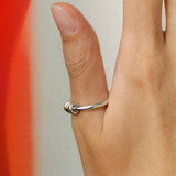 A close-up shows a hand wearing the SPINELLI KILCOLLIN Sirius 3 Annulets Ring—a minimalist sterling silver piece—on the middle finger, set against a red and beige gradient background to highlight its unisex design.