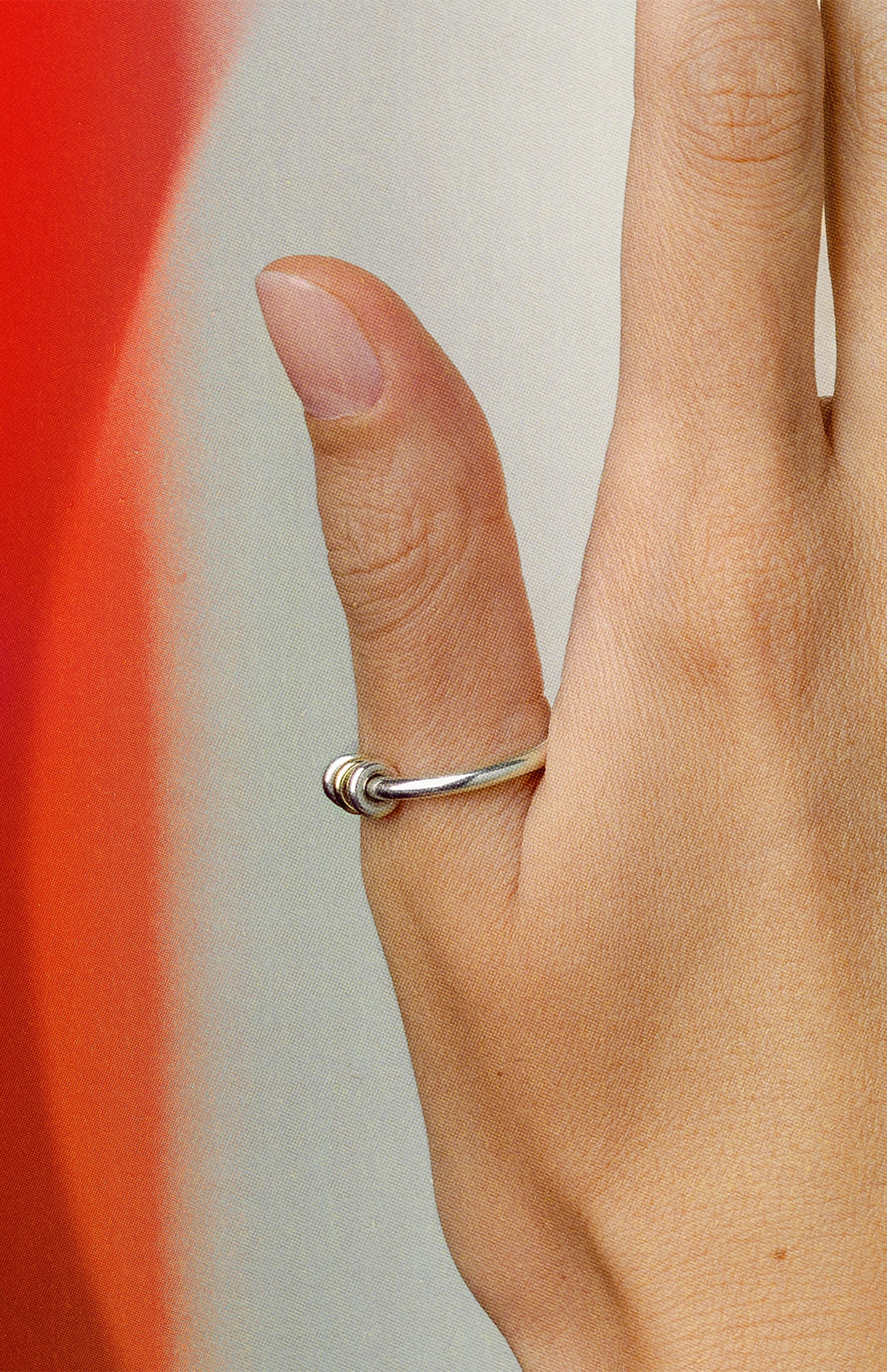 A close-up shows a hand wearing the SPINELLI KILCOLLIN Sirius 3 Annulets Ring—a minimalist sterling silver piece—on the middle finger, set against a red and beige gradient background to highlight its unisex design.