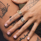 A close-up of tattooed hands with black nail polish, featuring butterfly and web designs. The fingers display the Solarium SG 3 Band Ring by SPINELLI KILCOLLIN, merging bold style with minimalism.