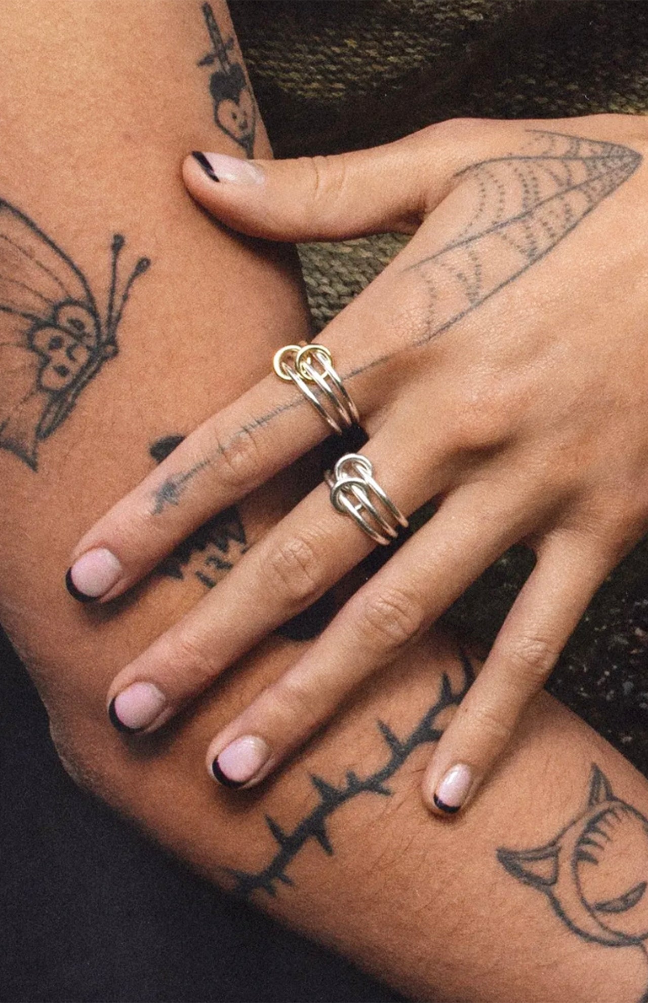 A close-up of tattooed hands with black nail polish, featuring butterfly and web designs. The fingers display the Solarium SG 3 Band Ring by SPINELLI KILCOLLIN, merging bold style with minimalism.