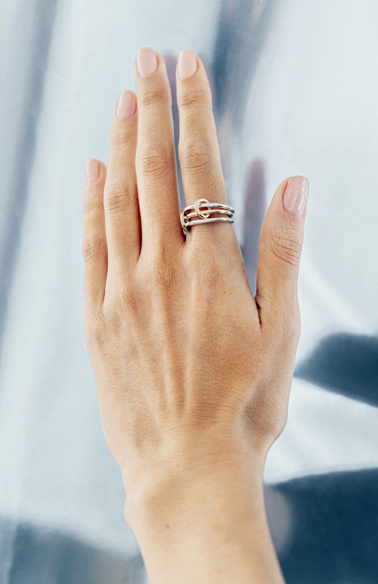 A hand with manicured nails is shown against a metallic background, wearing the SPINELLI KILCOLLIN Solarium 3 Band Ring in sterling silver and yellow gold, one band featuring a delicate knot for added elegance.