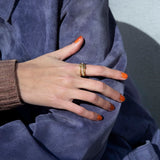 A person in a blue suede jacket and brown knitted sweater crosses their arm, displaying orange-painted nails and a SPINELLI KILCOLLIN Sonny Pavé Band Ring on their finger.
