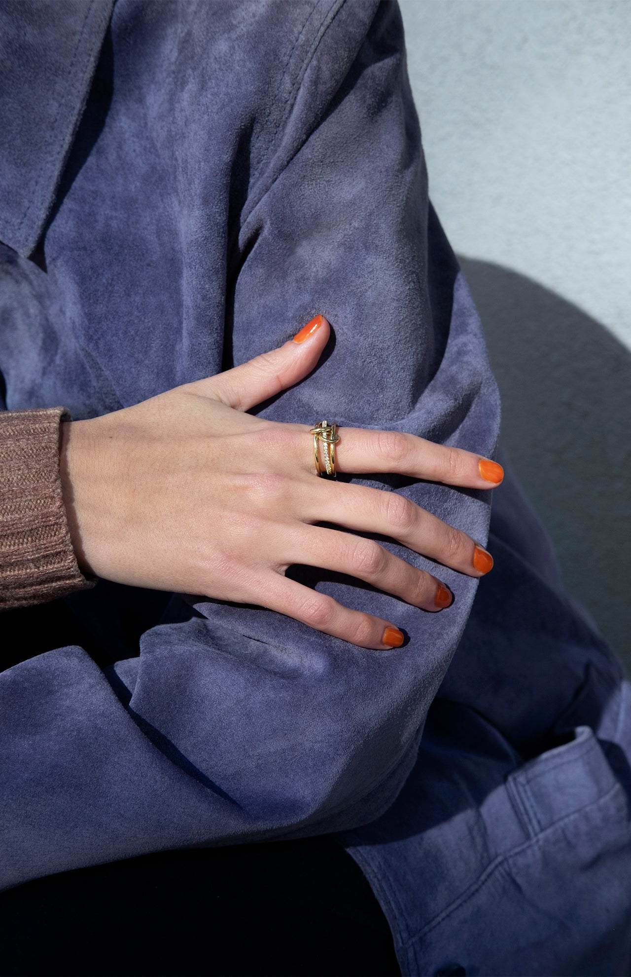 A person in a blue suede jacket and brown knitted sweater crosses their arm, displaying orange-painted nails and a SPINELLI KILCOLLIN Sonny Pavé Band Ring on their finger.