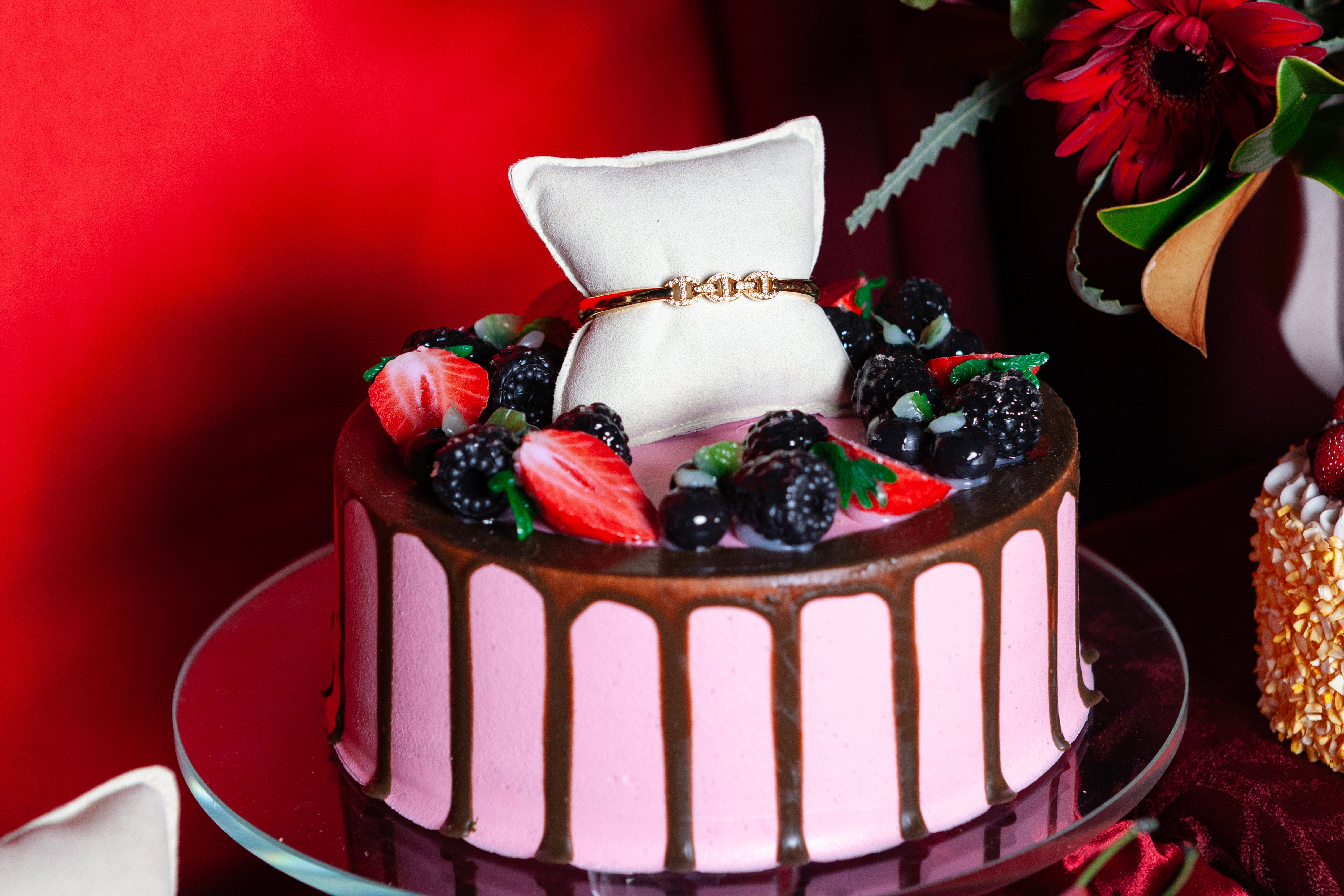 Chocolate cake with pink frosting and berries, featuring a ring box on top, against a red background.