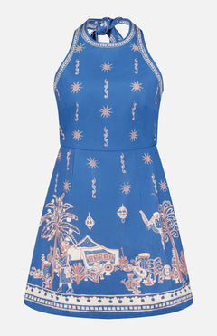 The Acacia Mini Halter Dress by ALEMAIS is a sleeveless blue organic cotton mini dress, adorned with white and tan palm trees, animals, houses, and decorative motifs along the hem and bodice.