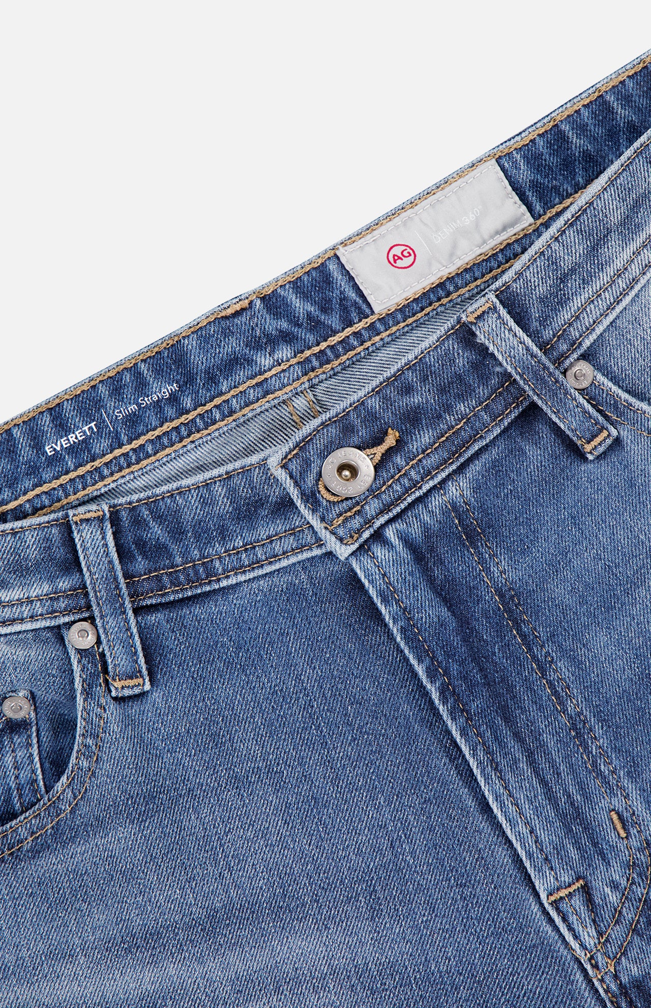 Close-up of AG Everett All-direction Stretch Jeans in stretch denim, showing metal button, zipper, belt loops, and front pockets. A white label with red AG logo appears inside the waistband.