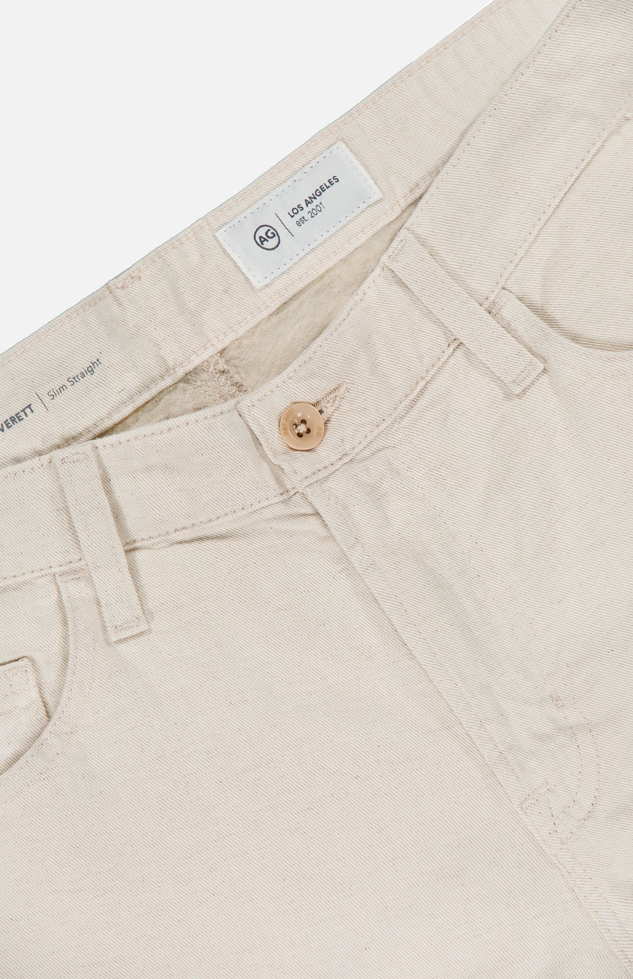 Close-up of the waistband and front of AG Everett Pants in light beige sustainable denim, featuring a wooden button, belt loops, front pockets, and an inner white label with AG branding and size details.