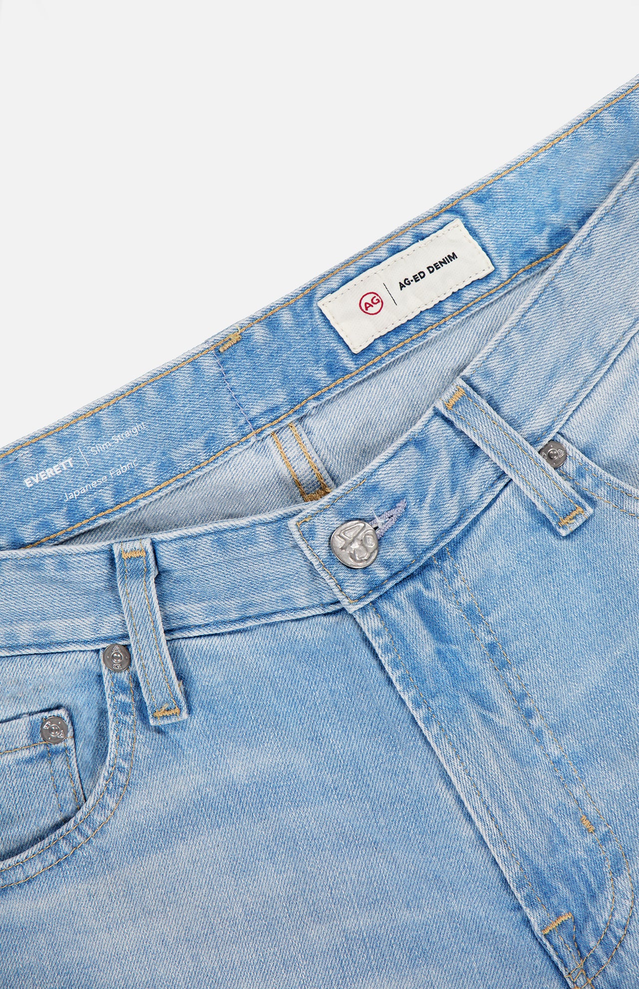 Close-up of the waistband and front of AG's Everett Jeans in 22 Years, a vintage-inspired slim fit style in light blue with belt loops, metal button, zipper, and two inside waistband labels including one reading A.B-D DENIM.