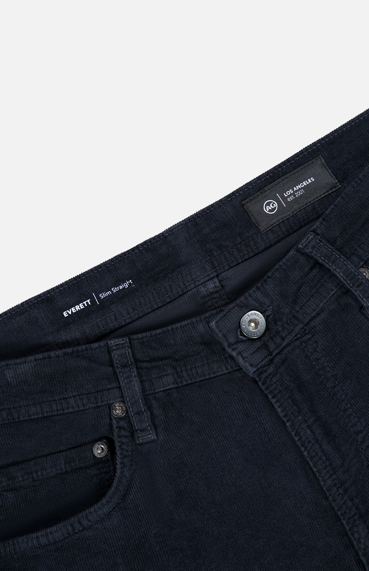 Close-up of the waistband and upper front section of the AG Everett Slim Corduroy Pant in dark navy, highlighting the buttons, pockets, belt loops, and an interior label displaying “LOS ANGELES” and “EVERETT Slim Straight.” This classic straight-leg design from AG lends a modern edge to your everyday style.