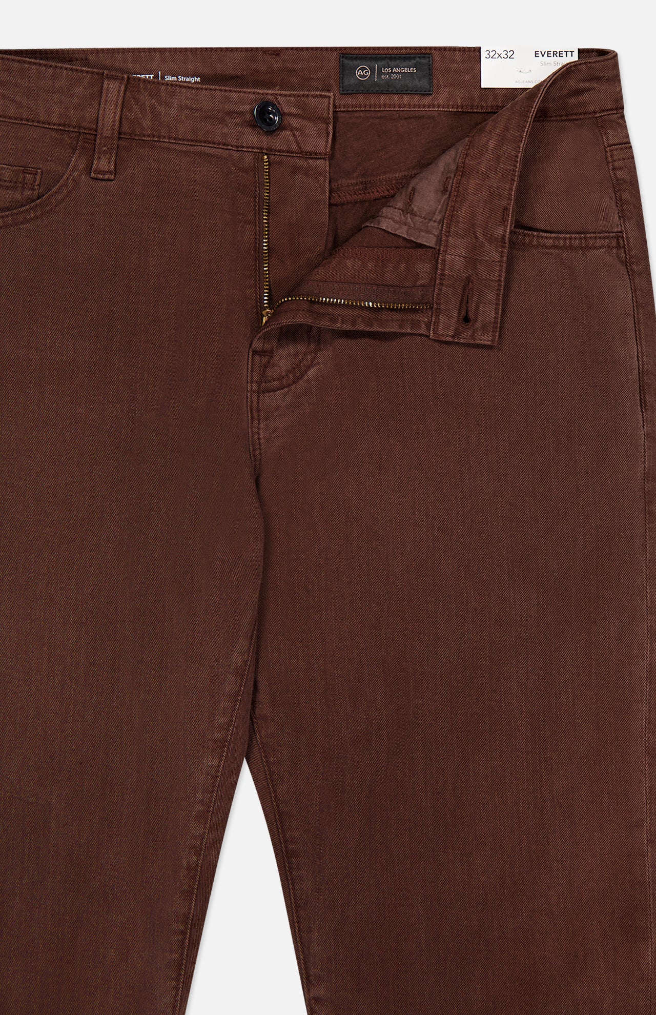 Close-up of AG’s Everett Pants in Sulfur, brown denim with the zipper and button undone, revealing the waistband and inside tag marked 32x32 slim fit.