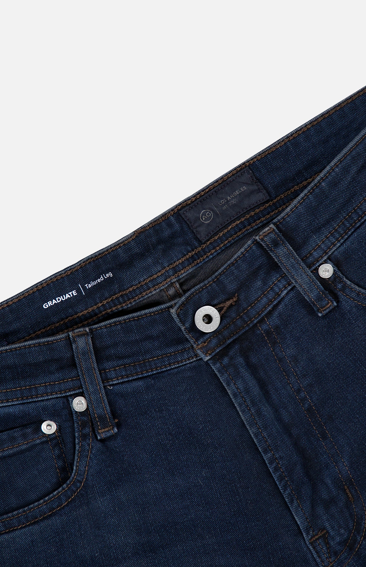 Close-up of the waistband and upper portion of the AG Graduate Pant in Studio, featuring a button closure, zipper fly, belt loops, and visible stitching on the pockets. The interior label reads GRADUATE | Tailored Leg, emphasizing vintage-inspired detailing throughout the design.