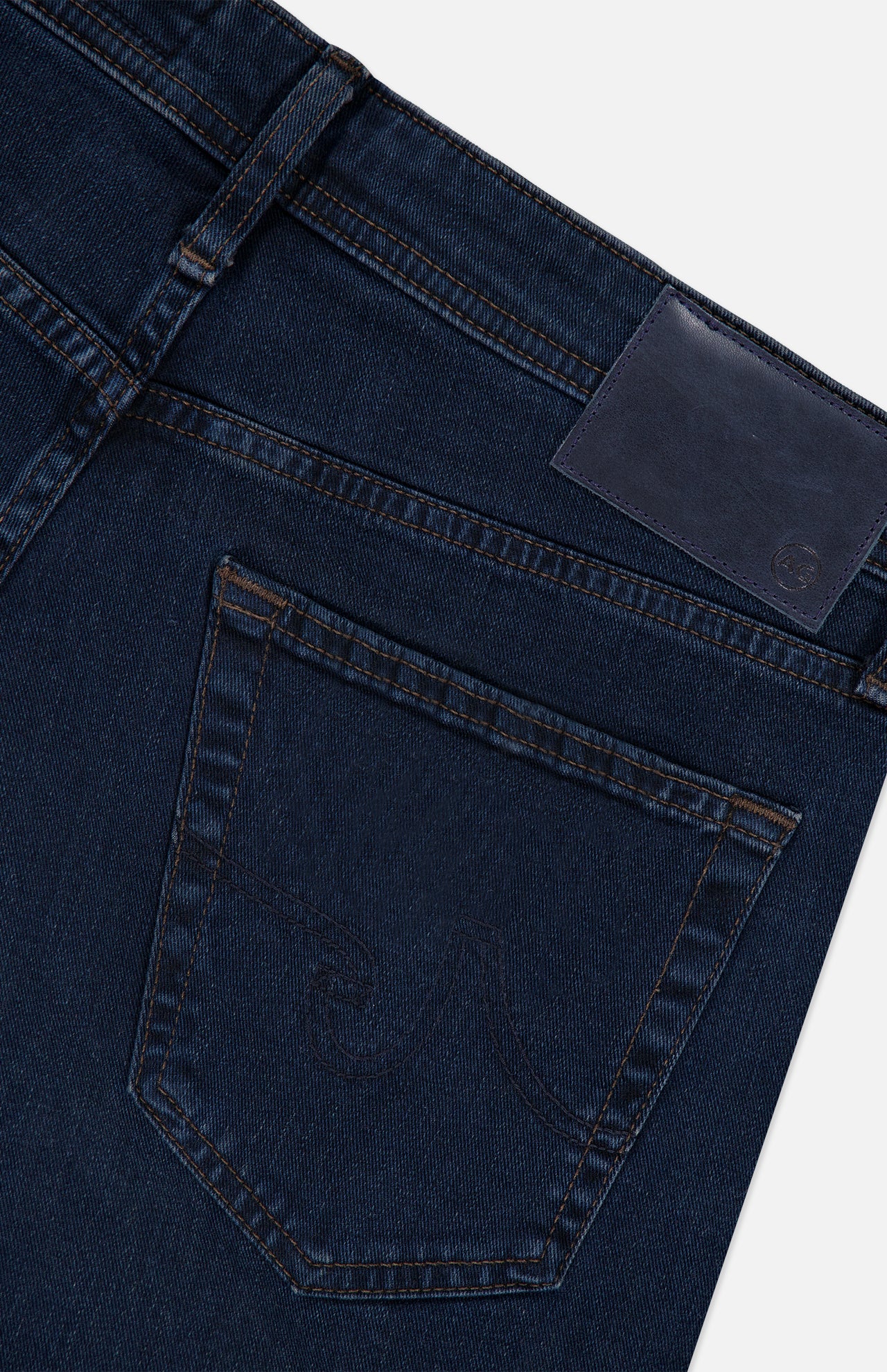 Close-up of the back pocket and waistband of the AG Graduate Pant in Studio, featuring ultra-dark blue denim with visible stitching, vintage detailing, and a blank leather patch on the waistband.