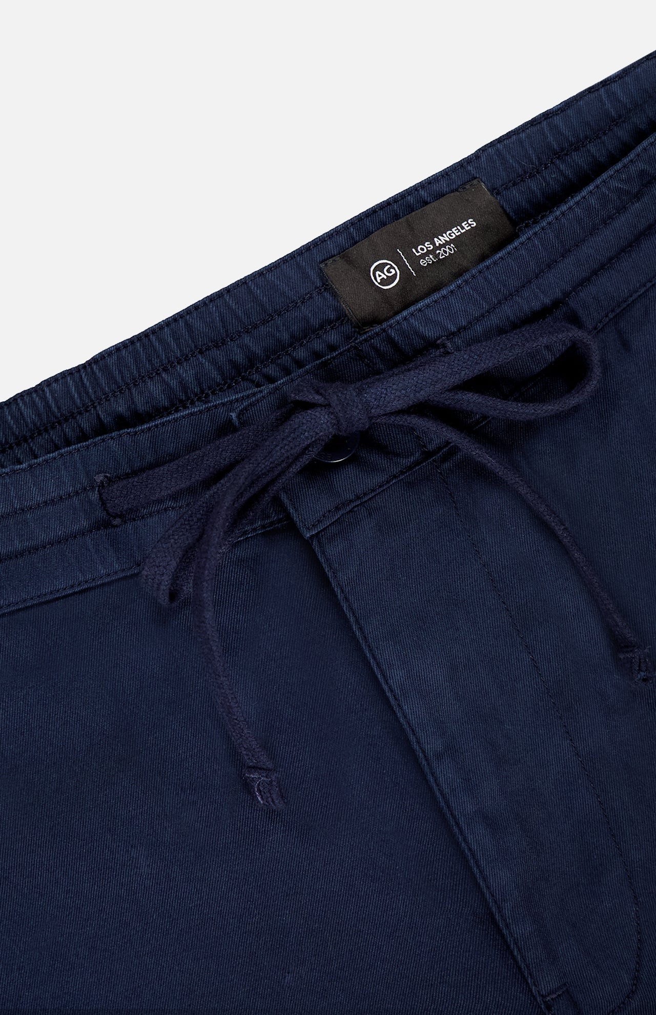 Close-up of AG Oliver Drawstring Shorts in navy blue, featuring a comfortable mid rise, elastic waistband with drawstring, and visible AG brand label. Fabric appears to be cotton or a cotton blend.