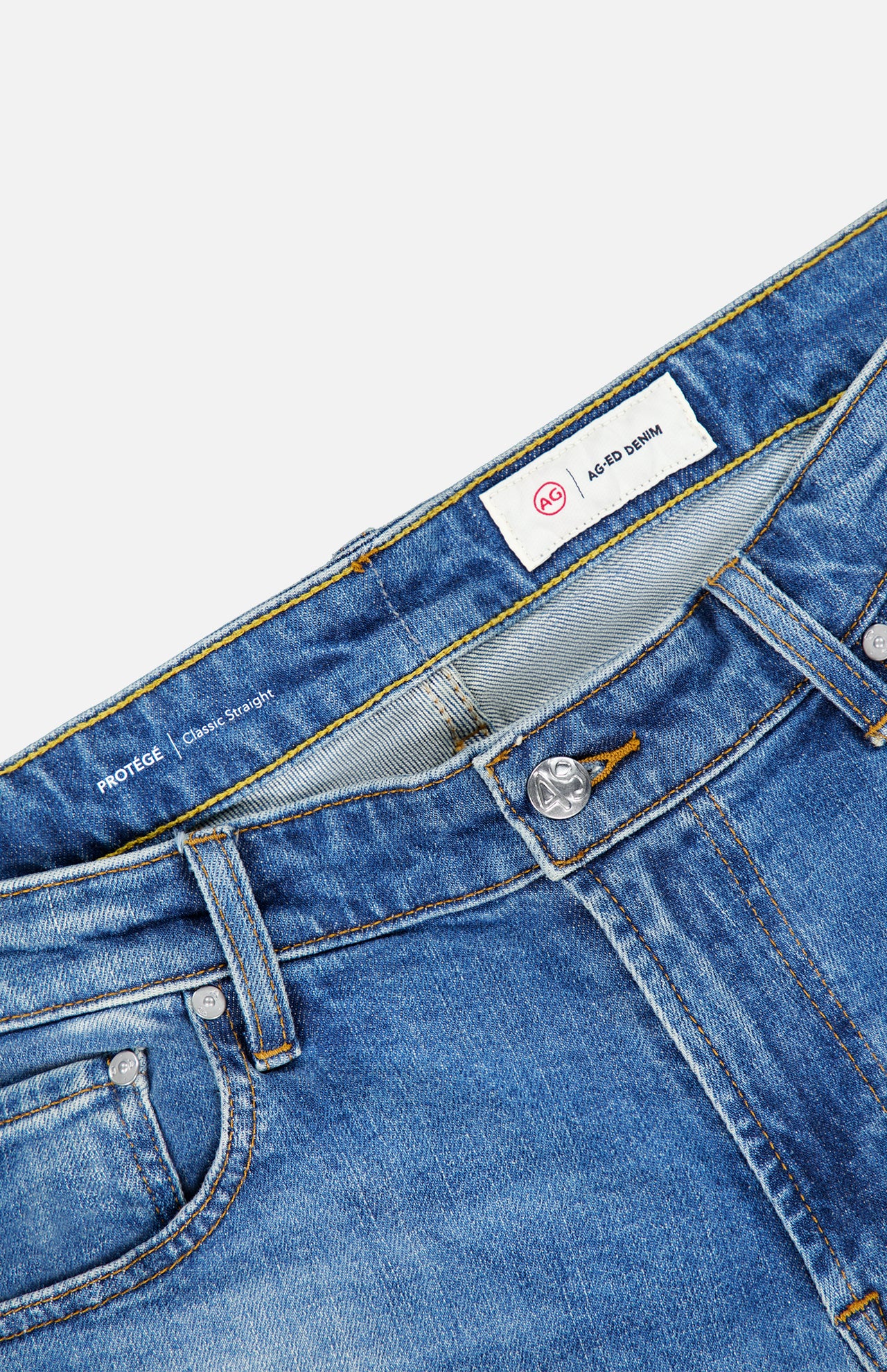 Close-up of the Protege Jean in 14 Years by AG, featuring blue denim with visible stitching, belt loops, a button closure, and a white label reading “AG AGED DENIM” inside the waistband. The straight leg fit is accentuated by subtle vintage details throughout the fabric.