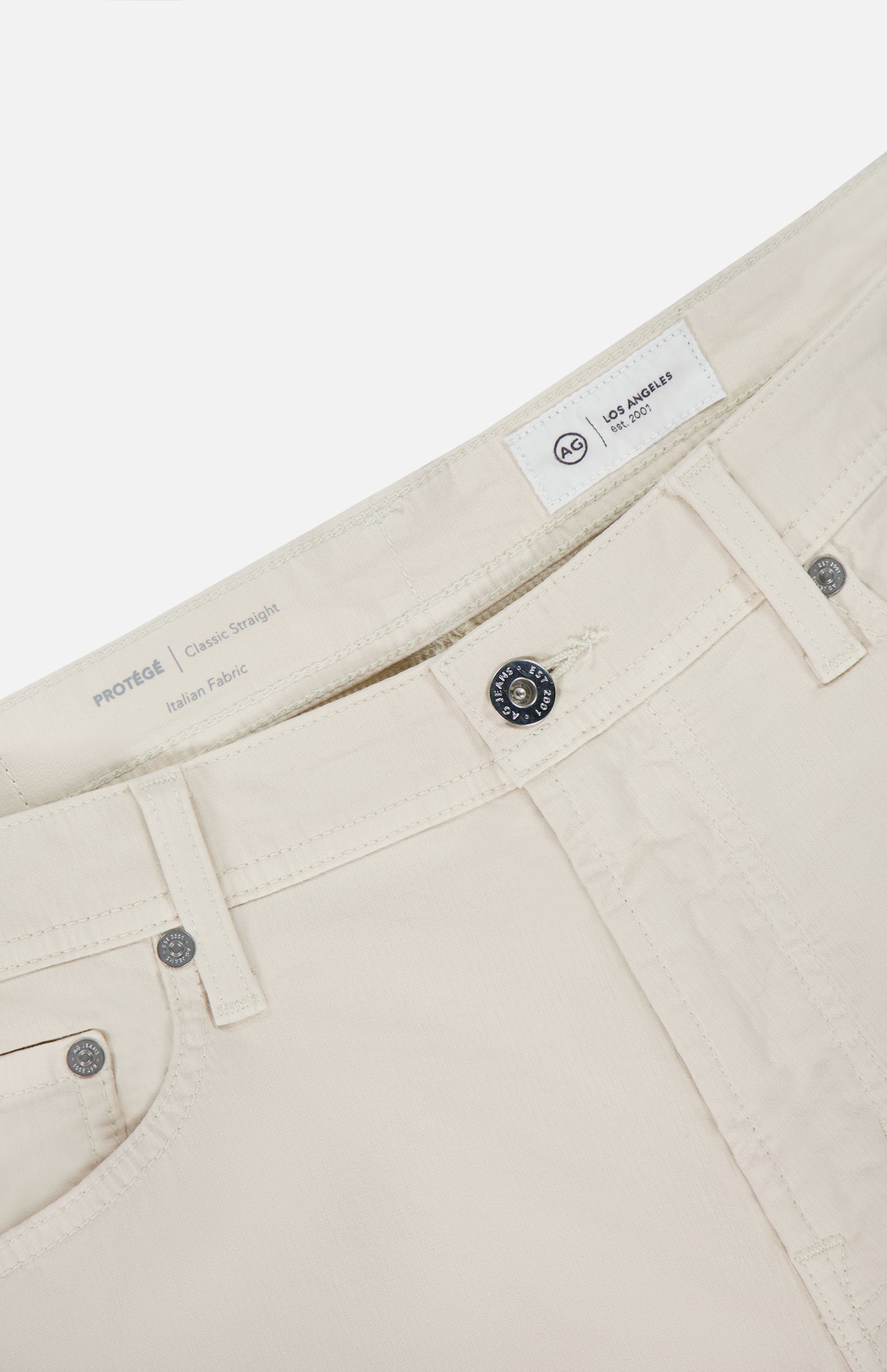 Close-up of the waistband and upper front of AG Protege Pants in Pumice, highlighting the button, zipper, belt loops, and interior tags with a label reading PROTÉGÉ Classic Straight Italian Fabric.