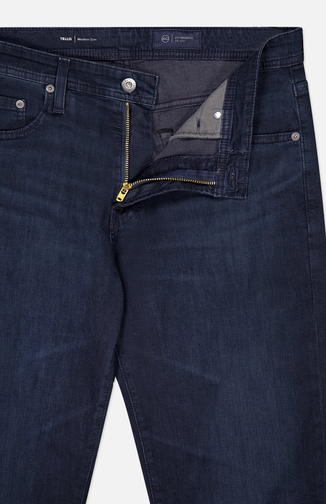 The AG Tellis Slim Jeans in Tamarack are dark blue with a slightly faded texture, classic pockets, and belt loops. Shown with the zipper and top button undone to reveal the inside waistband and fly area.