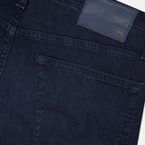 Close-up of the back of AG Tellis Slim Jeans in Tamarack, featuring dark blue denim, a subtly stitched back pocket, and a navy leather AG logo patch near the waistband.