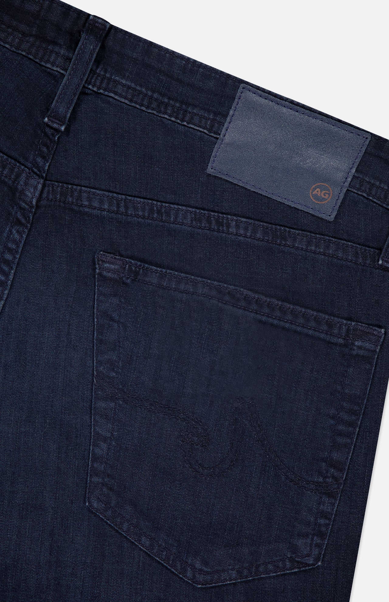 Close-up of the back of AG Tellis Slim Jeans in Tamarack, featuring dark blue denim, a subtly stitched back pocket, and a navy leather AG logo patch near the waistband.