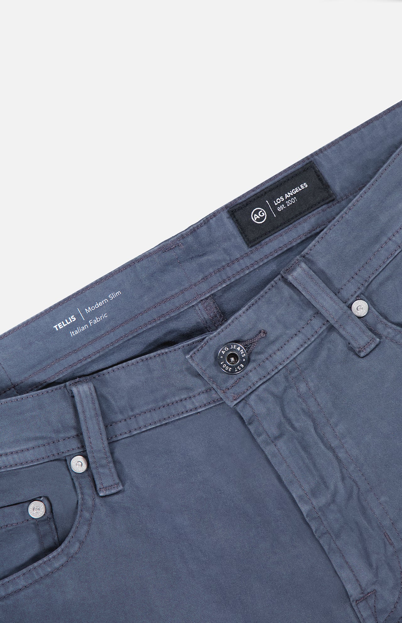 A close-up of the AG Tellis Sueded Sateen Pants in Blue Grain, featuring Italian stretch fabric with a microsueded finish, and details like the waistband, button, front pockets, and the Tellis Modern Slim Italian Fabric label inside.